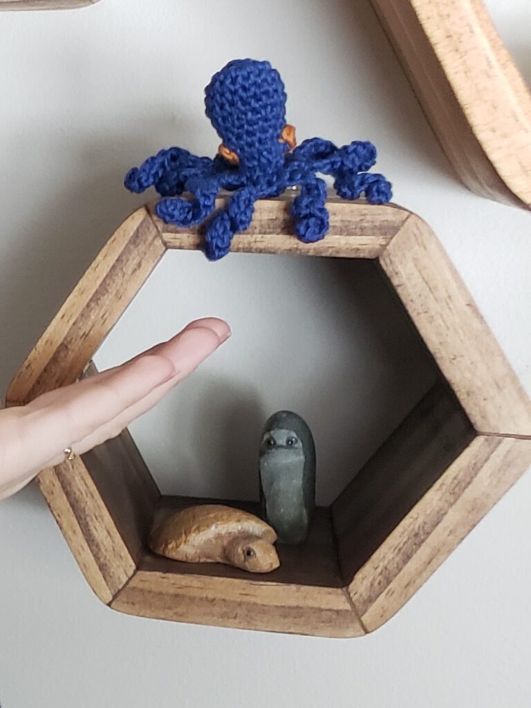 Tiny crocheted octopus on a shelf with a hand for reference